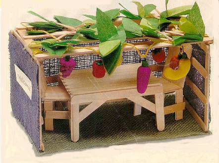 A craft sukkah, complete with walls, roof, table, chairs, and decorations, made of lolly sticks and tissue paper.