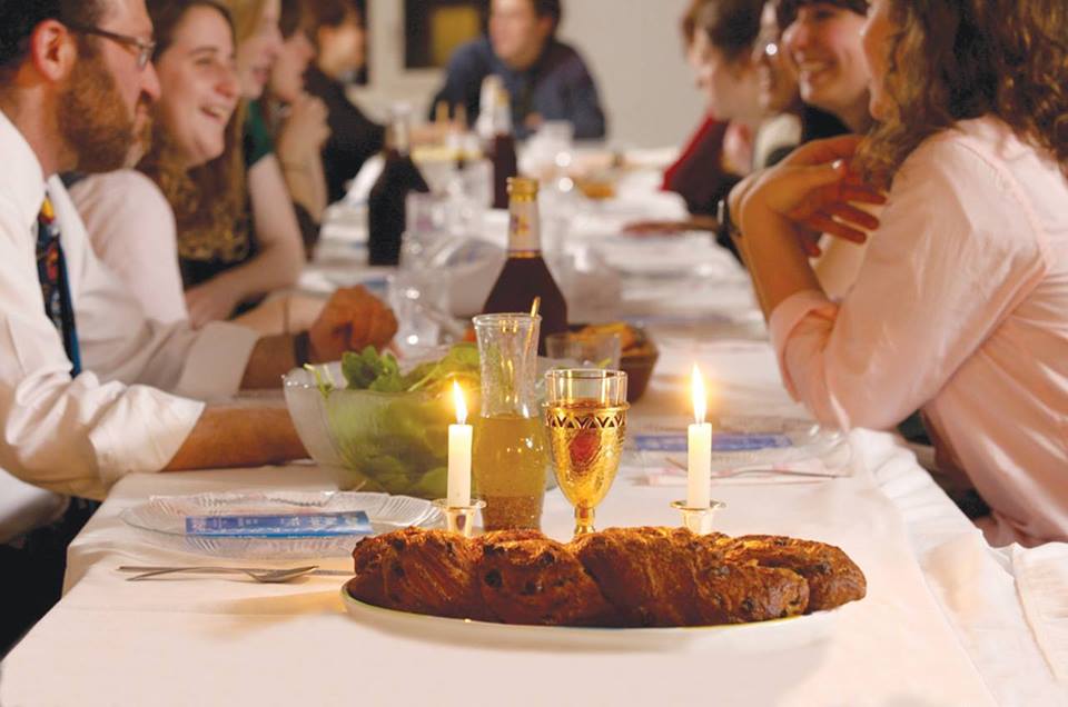 smiling people sit around a table that is set for Friday night dinner with wine and Shabbat candles