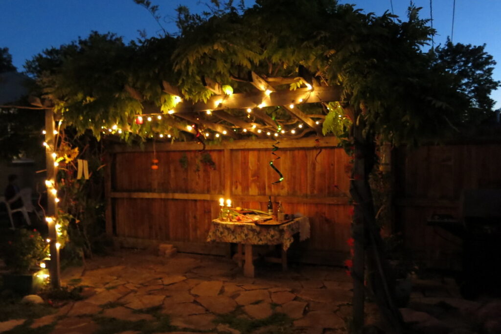 View into a decorated Sukkah against a night sky. Inside the sukkah there is a table with lit candles, a lulav and etrog, and wine.